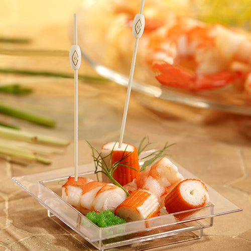 Su un piatto quadrato di plastica con imitazione di granchio, gamberetti, uova verdi e guarnizione, sono raffigurati due PAPSTAR 40 spiedini finger food "pure" della PAPSTAR GmbH, sullo sfondo si vede sfocato un piatto simile di frutti di mare.