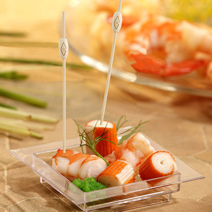 Su un piatto quadrato di plastica con imitazione di granchio, gamberetti, uova verdi e guarnizione, sono raffigurati due PAPSTAR 40 spiedini finger food "pure" della PAPSTAR GmbH, sullo sfondo si vede sfocato un piatto simile di frutti di mare.