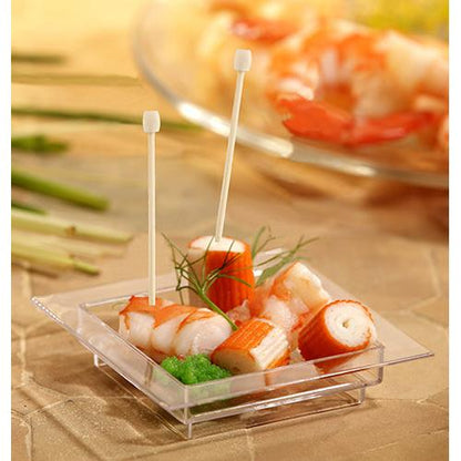 Una piccola ciotola quadrata trasparente contiene surimi, guarnizione verde e due PAPSTAR 100 Fingerfood - Picker di PAPSTAR GmbH. Sullo sfondo appaiono verdure sfocate e altri frutti di mare.
