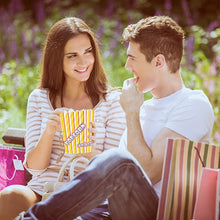 Una donna e un uomo siedono sorridendo su una panchina all'aperto e condividono popcorn dai PAPSTAR 100 sacchetti di popcorn, sostituto della pergamena "Popcorn" resistente al grasso della PAPSTAR GmbH, nelle vicinanze ci sono sacchetti della spesa.