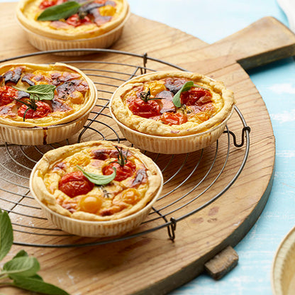 Tre piccoli quiche di verdure con pomodorini e erbe si trovano su una griglia rotonda su un tagliere di legno, circondati da foglie di erbe fresche e PAPSTAR 50 stampi "pure" marroni della PAPSTAR GmbH.