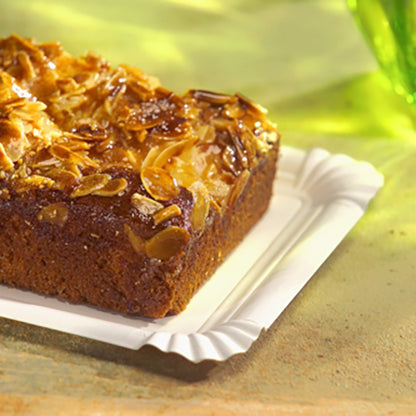 Sul PAPSTAR piatto di cartone rettangolare 13x20 cm, bianco, "pure" per feste (12 pezzi) della PAPSTAR GmbH riposa una torta rettangolare con mandorle a scaglie - un'opzione di servizio ecologica ed elegante su una superficie chiara con vetro verde dietro.