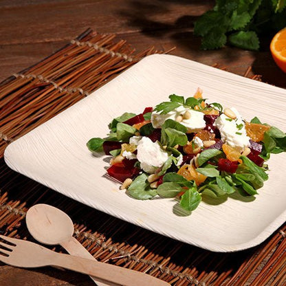 Un PAPSTAR piatto quadrato in foglia di palma "pure" da 6 della PAPSTAR GmbH, contiene un'insalata verde mista con barbabietola, fette d'arancia, formaggio di capra e noci. È posizionato su un tappetino di bambù con posate di legno; sullo sfondo si vedono un'arancia e delle erbe.
