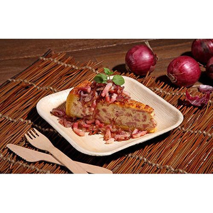 Un pezzo di tortilla spagnola con cipolle rosse viene servito su un piatto quadrato "Pure" della PAPSTAR GmbH dal set da 6 PAPSTAR, accompagnato da coltello e forchetta di bambù. Sullo sfondo si vedono cipolle rosse con la buccia su un tappetino intrecciato.