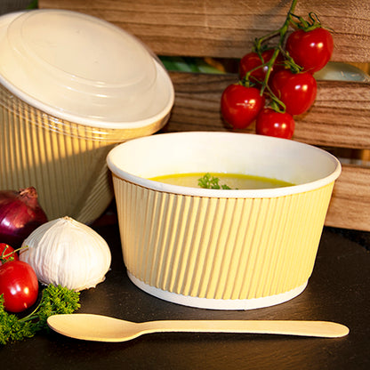 Un PAPSTAR bicchiere da zuppa rotondo, marrone/bianco, Ripple Wall (13,5 cm), riempito con zuppa, si trova accanto a un cucchiaio di legno su una superficie scura. Nelle vicinanze ci sono pomodori a grappolo, aglio, cipolla, prezzemolo e un altro bicchiere di carta della PAPSTAR GmbH con coperchio.