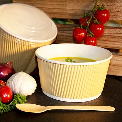 Un bicchiere da zuppa Starpak giallo con zuppa e guarnizione di erbe è posizionato accanto a un cucchiaio di legno, aglio, cipolle rosse, pomodorini e prezzemolo; sullo sfondo si vede un coperchio rotondo Starpak 25 per bicchieri da zuppa R-PET.
