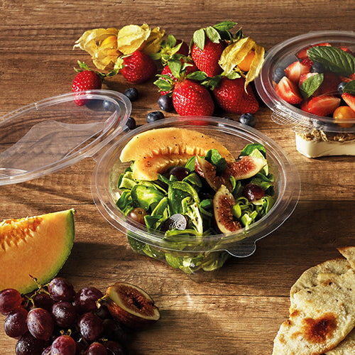 Su un tavolo di legno c'è una ciotola rotonda Starpak 50 per gastronomia e insalata con coperchio a cerniera (R-PET trasparente) con insalata di melone, fichi e verdure a foglia. Accanto ci sono fragole, uva, fette di melone, pomodorini, pane pita e un'altra ciotola con pomodori e basilico.