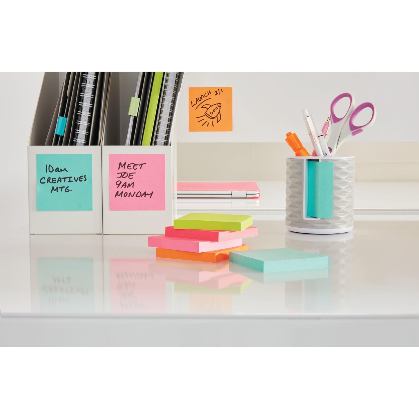 Una scrivania ordinata con cubi colorati di Post-it® Notes (76x76mm, 450 fogli per blocco, 100% PEFC) della 3M Deutschland GmbH, un portapenne bianco pieno di penne e forbici, quaderni e scatole di schede. Le note adesive sono contrassegnate da promemoria scritti a mano.