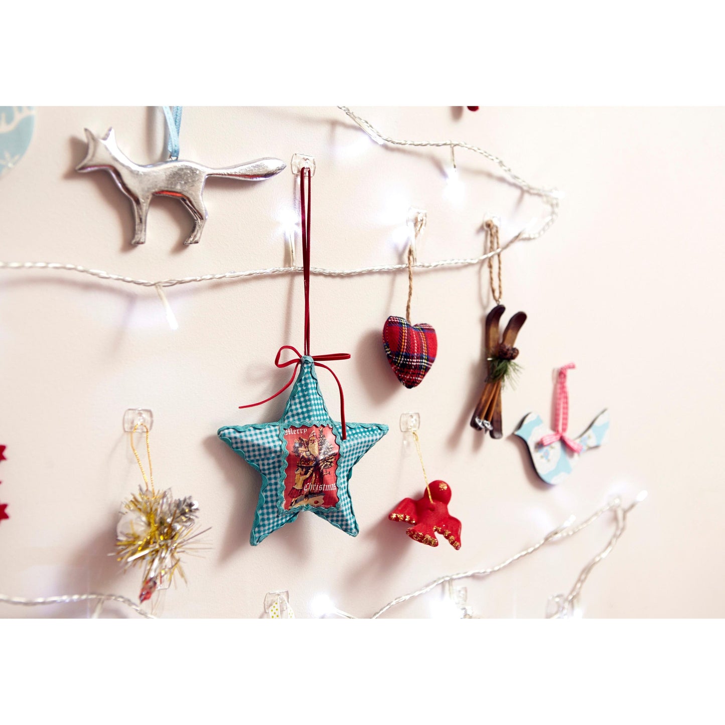 An einer beigen Wand hängen weiße Lichterketten und festliche Ornamente - darunter ein silberner Fuchs, ein blauer Stern, ein kariertes Herz und eine weiße Taube - an transparenten Command™ Dekorations-Clips der 3M Deutschland GmbH.