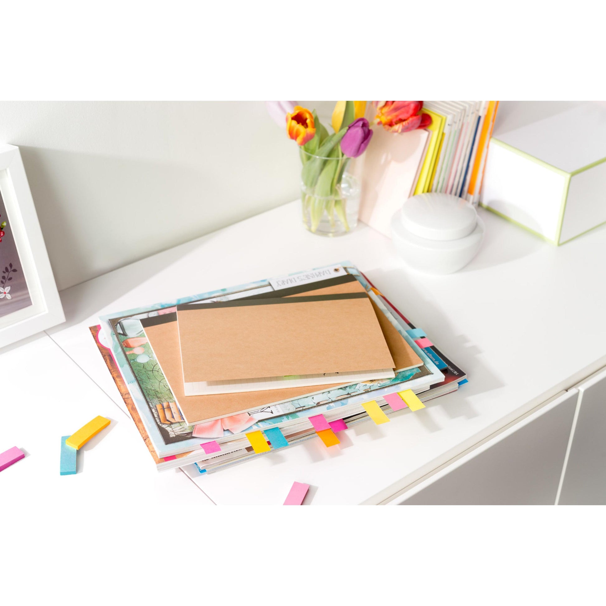 Ein Stapel Bücher und Hefte mit Post-it® Page Markern (15 mm x 50 mm, 100 Blatt/Block, 5 Blöcke/Packung) der 3M Deutschland GmbH in verschiedenen Farben liegt auf einem weißen Schreibtisch. Lose Marker liegen verstreut herum.