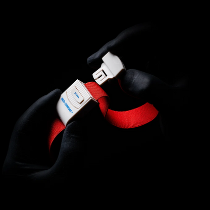 Mani guantate che ricordano un laccio emostatico AMPri MED-COMFORT afferrano la fibbia di una cintura di sicurezza rosso brillante davanti a uno sfondo ombreggiato. Sulla fibbia si leggono la parola "Blue" e la lettera "B".