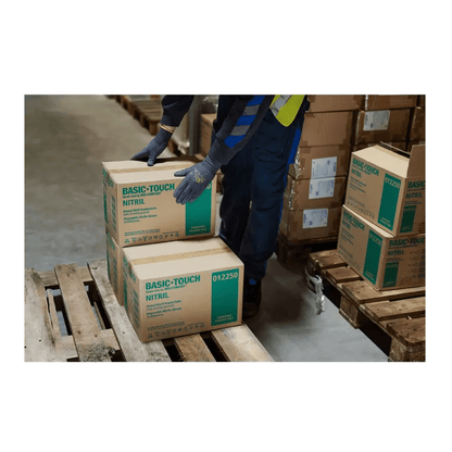 Una persona che indossa guanti da montaggio ultraleggeri AMPri SolidSafety Tough Micro Touch impila accuratamente scatole con la scritta "Basic-Touch Nitrile" su un pallet di legno in un magazzino. Diverse altre scatole sono visibili e la persona sembra ordinare il magazzino con precisione e cura.