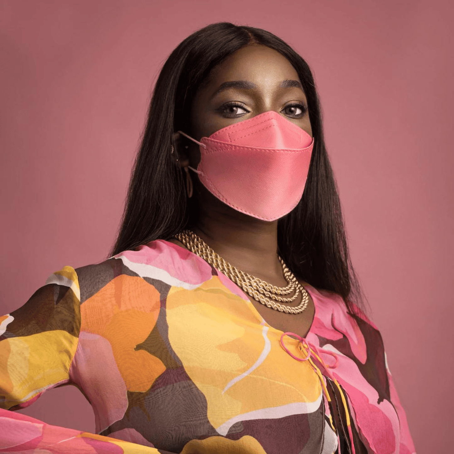 Una persona indossa una maschera facciale designer Altruan filtrante particelle (standard FFP2) in un elegante rosa e una maglietta colorata con un motivo astratto su uno sfondo rosa. Ha i capelli lunghi e scuri e indossa diverse collane d'oro.