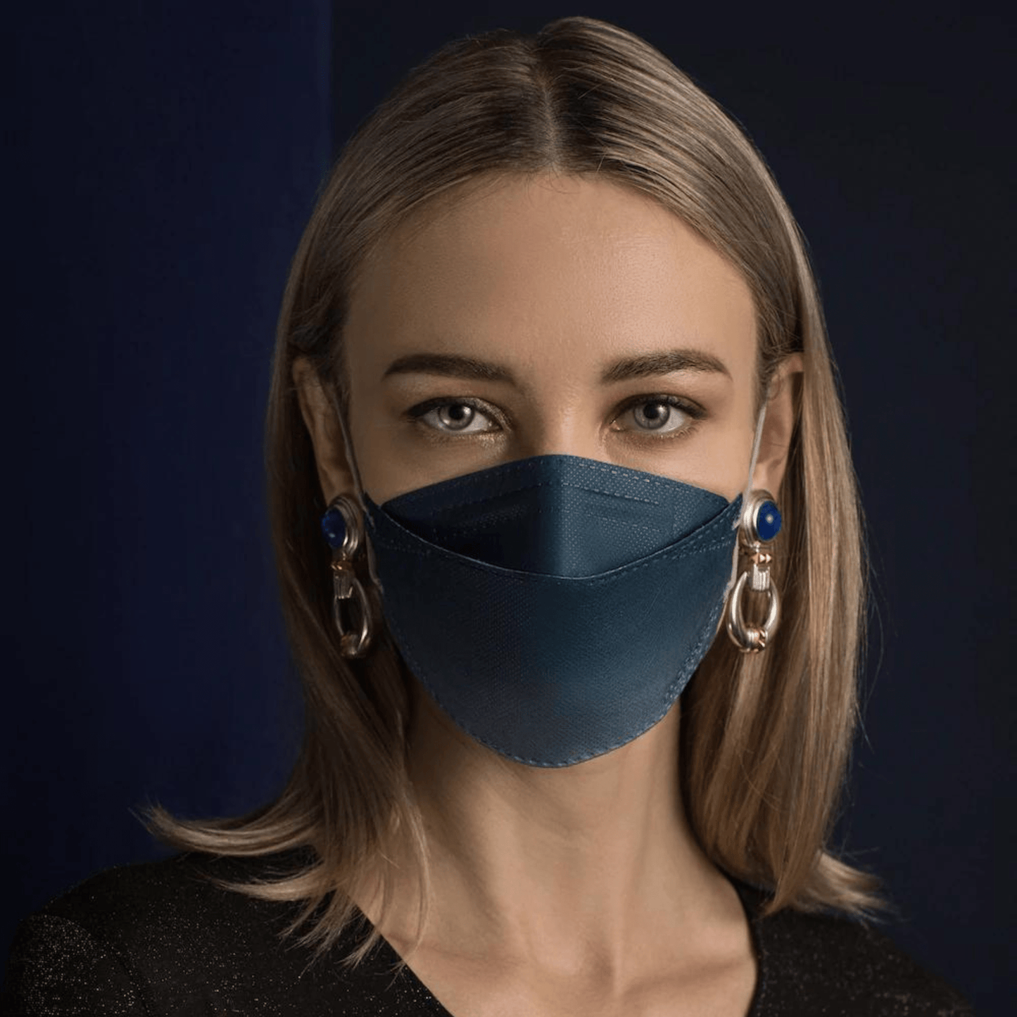 Una persona con i capelli lunghi indossa una maschera facciale designer Altruan filtrante particelle in blu, che offre una protezione efficace secondo lo standard FFP2. La abbina a grandi orecchini decorativi su uno sfondo scuro e indossa una camicia nera.