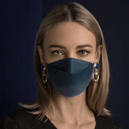 Una persona con i capelli lunghi indossa una maschera facciale designer Altruan filtrante particelle in blu, che offre una protezione efficace secondo lo standard FFP2. La abbina a grandi orecchini decorativi su uno sfondo scuro e indossa una camicia nera.