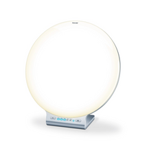 Una lampada diurna TL 100 rotonda e bianca Beurer della Beurer GmbH su un supporto argentato con un piccolo pannello di controllo nella parte inferiore con pulsanti e un display digitale. La lampada emette una luce morbida e uniforme, perfetta come luce d'atmosfera, con il marchio „Beurer“ stampato in alto al centro.