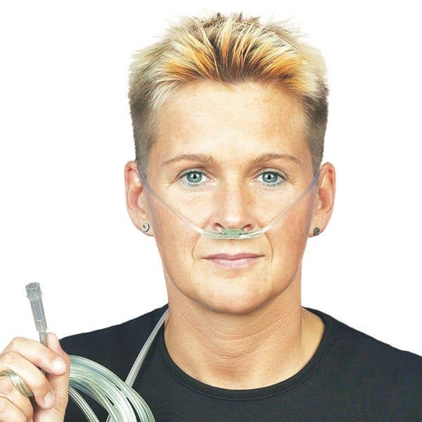 Eine Person mit kurzen Haaren verwendet die DCT Sauerstoffbrille Soft für Erwachsene der Servoprax GmbH zur Sauerstoffunterstützung. Sie hält den O2-Verbindungsschlauch und trägt ein schwarzes Hemd vor einem schlichten weißen Hintergrund.