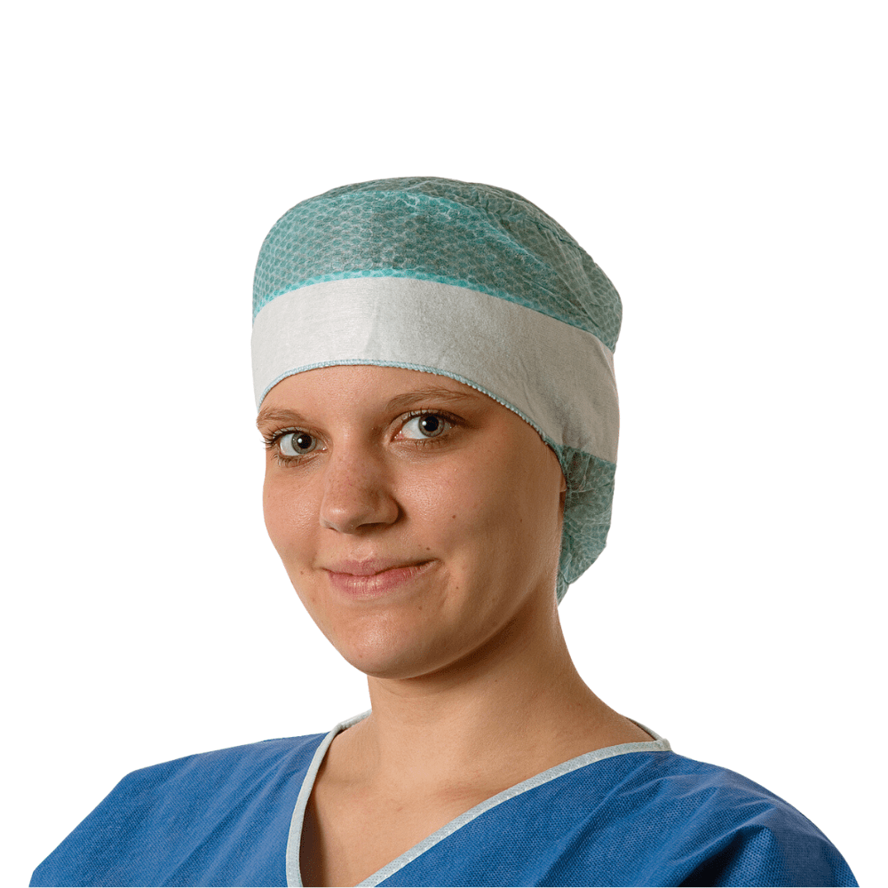 Una giovane dottoressa indossa un camice chirurgico blu e un cappello monouso Hartmann Foliodress® Cap Comfort Form Plus in tessuto non tessuto viscosa traspirante e sorride dolcemente alla fotocamera. Lo sfondo è semplicemente bianco.