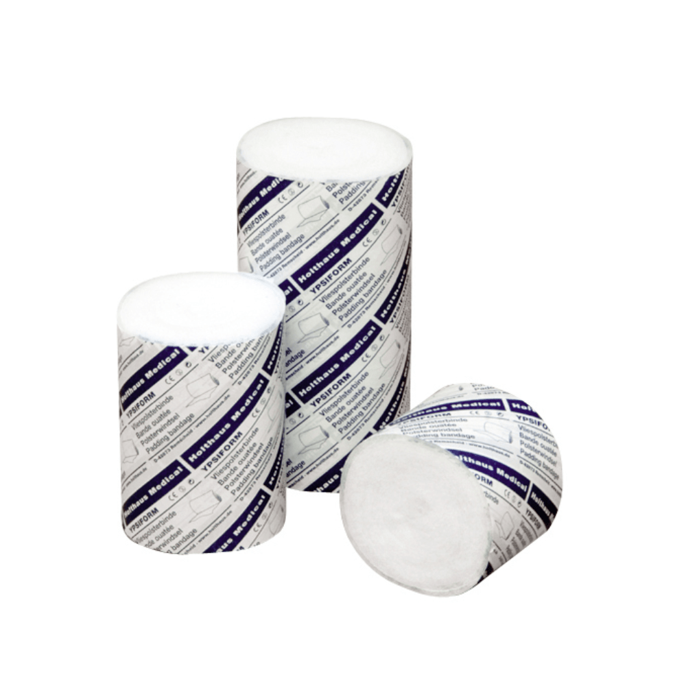 Sono mostrati tre rotoli cilindrici di cotone in confezioni bianche e blu parzialmente aperte. Due rotoli di Holthaus Ypsiform Polsterbinde della Holthaus Medical GmbH & Co. KG sono in posizione verticale, mentre un rotolo è sdraiato e mostra l'estremità di cotone bianco.