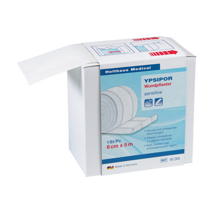 È raffigurata una scatola di cerotti Holthaus Ypsipor in tessuto non tessuto della Holthaus Medical GmbH & Co. KG. Sulla scatola è indicato che è delicato, misura 6 cm x 5 m ed è prodotto in Germania. Una parte del rotolo di cerotto in tessuto non tessuto delicato sulla pelle è visibile e sporge dalla scatola.