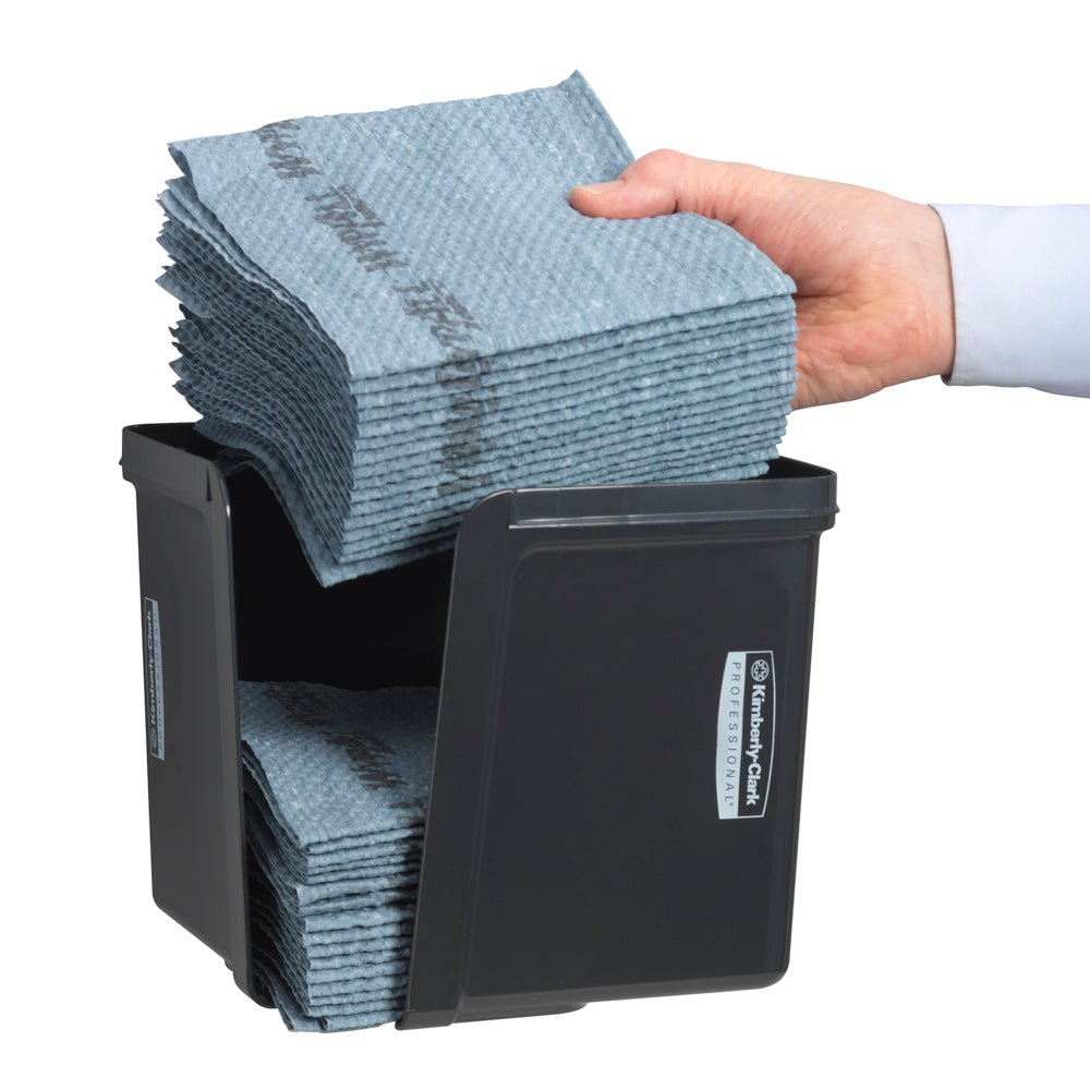 Una mano inserisce salviette piegate in quarti nel distributore di salviette Kimberly-Clark Professional™ grigio di Kimberly-Clark GmbH - per un'estrazione igienica dei panni. I panni ordinatamente piegati e l'etichetta anteriore sono visibili.