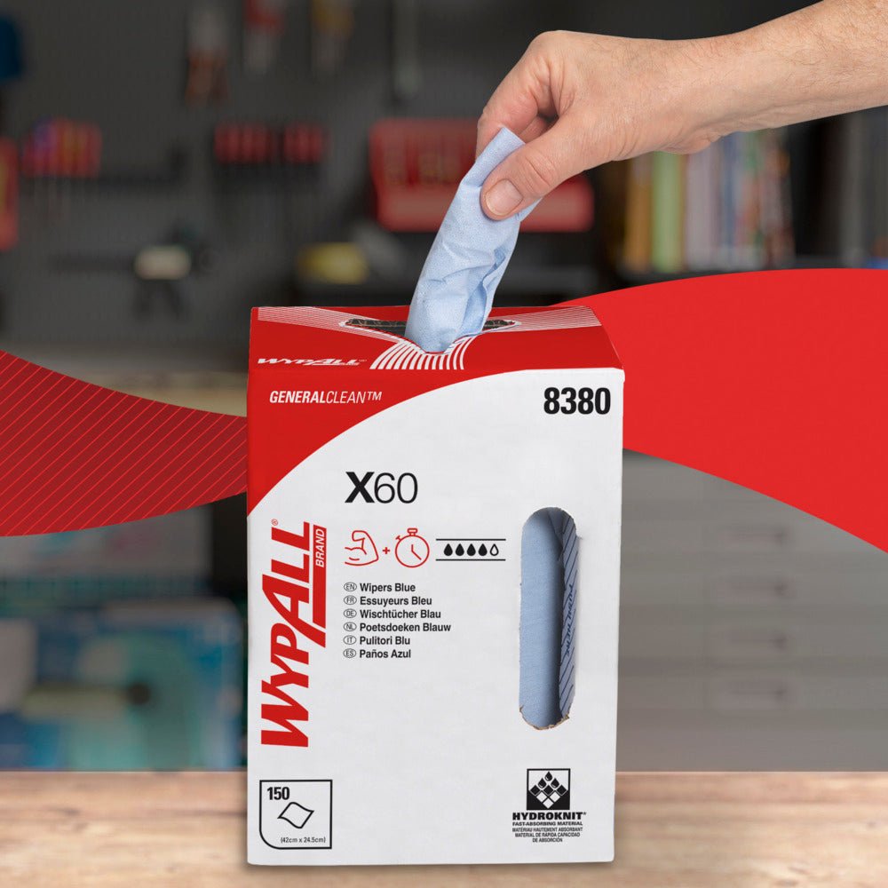 Una mano estrae un foglio blu di WypAll® X60 General Clean da una scatola dispenser bianco-rossa della Kimberly-Clark GmbH con la scritta "8380", "150 fogli" e "Estrazione centrale". Sullo sfondo si vede un'officina sfocata con scaffali.