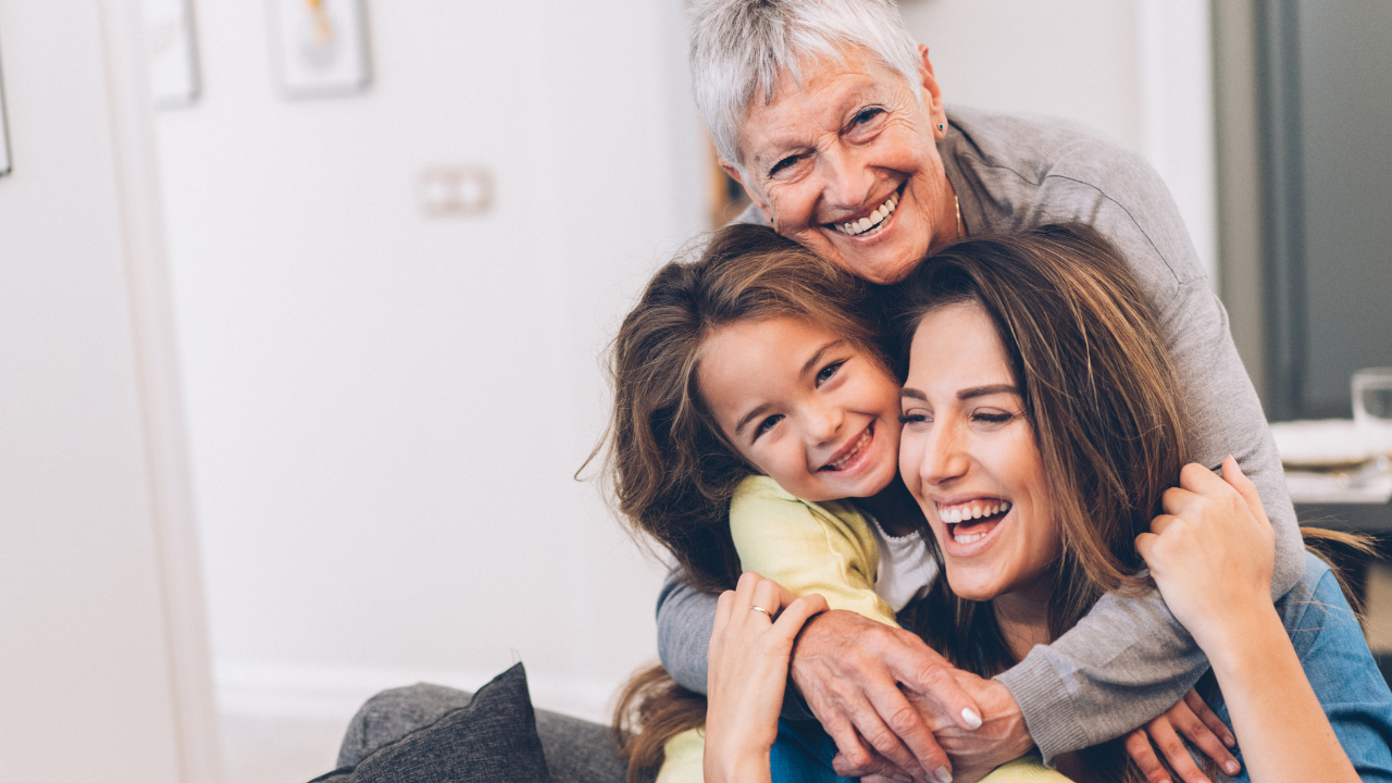 Das Bild zeigt drei Generationen von Frauen – eine ältere Dame, eine Mutter und ihre Tochter. Alle drei sind in einem hellen, freundlichen Raum zu sehen und umarmen sich glücklich. Die ältere Dame lacht und ist hinter der Mutter und Tochter, die ebenfalls lächeln und sich in der Mitte des Bildes befinden. Die Atmosphäre vermittelt Liebe, Freude und familiäre Nähe. Es scheint ein besonderer Moment des Zusammenseins und der Zuneigung zu sein.