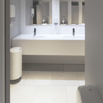 Si vede un bagno pubblico pulito e moderno con un lungo piano bianco con due lavandini e rubinetti. Sotto il piano spicca una parete di piastrelle bianche. La stanza è dotata di specchi, dispenser di sapone Tork di Essity Professional Hygiene Germany GmbH e un cestino bianco su un pavimento piastrellato, progettato per bagni ad alto traffico.