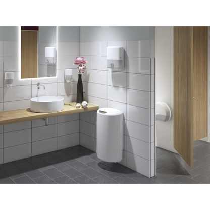 Un bagno pubblico moderno con pareti piastrellate bianche e pavimento grigio scuro è un punto forte del design Elevation. Dispone di una mensola in legno con lavandino rotondo, grande specchio, distributore Tork 555000 per carta igienica Mini-Jumbo Elevation T2 di Essity Professional Hygiene Germany GmbH, nonché un distributore di sapone, distributore di asciugamani, vaso di fiori, cestino per i rifiuti e una porta della cabina parzialmente aperta.