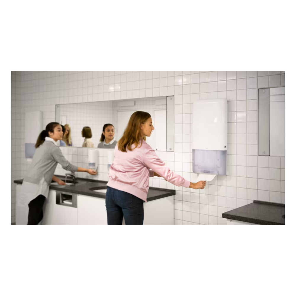 Due donne si trovano in un bagno con pareti piastrellate bianche e grandi specchi. Una utilizza un lavandino sul lato sinistro, mentre l'altra estrae senza sforzo un asciugamano infinito da un distributore Tork PeakServe® 552500 per asciugamani infiniti Elevation H5 di TORK - Essity Professional Hygiene Germany GmbH, situato sul lato destro. Il piano del lavabo grigio completa l'illuminazione luminosa in questo bagno molto frequentato.