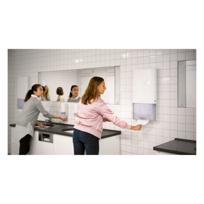 Due donne si trovano in un bagno con pareti piastrellate bianche e grandi specchi. Una utilizza un lavandino sul lato sinistro, mentre l'altra estrae senza sforzo un asciugamano infinito da un distributore Tork PeakServe® 552500 per asciugamani infiniti Elevation H5 di TORK - Essity Professional Hygiene Germany GmbH, situato sul lato destro. Il piano del lavabo grigio completa l'illuminazione luminosa in questo bagno molto frequentato.
