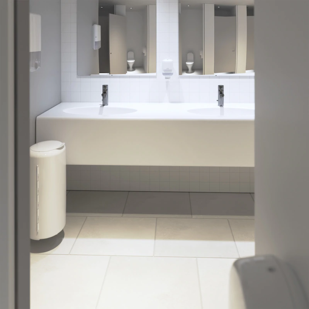 Il moderno bagno pubblico dispone di un elegante piano lavabo bianco con due lavandini e rubinetti cromati. A sinistra c'è un cestino bianco, mentre sopra il pavimento piastrellato sono appesi specchi. Sulla parete sono montati dispenser di sapone Tork Xpress® 552000 di Essity Professional Hygiene Germany GmbH, e gli asciugamani multifoglio aumentano il comfort dei visitatori.