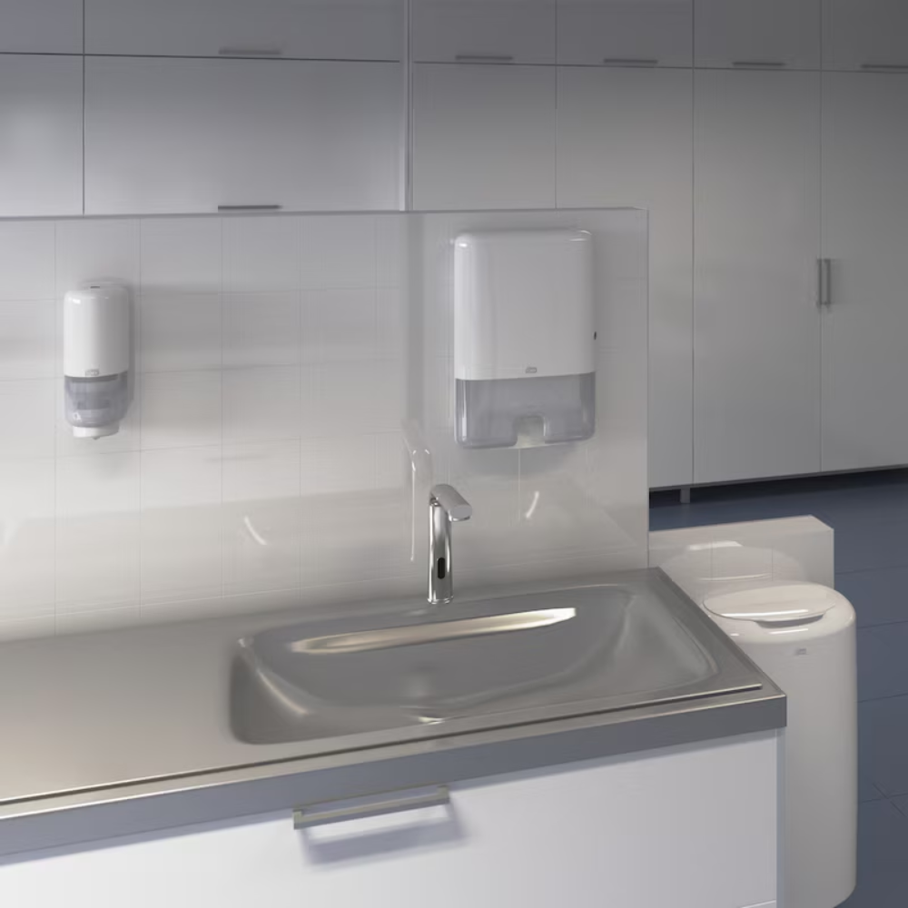 Viene mostrato un moderno lavandino in un bagno pubblico con rubinetto automatico. Sopra il lavandino sono montati su una parete piastrellata un dispenser Tork Xpress® 552000 per asciugamani multifoglio H2 di Essity Professional Hygiene Germany GmbH e un dispenser di sapone. Sullo sfondo si vede una fila di armadietti bianchi.