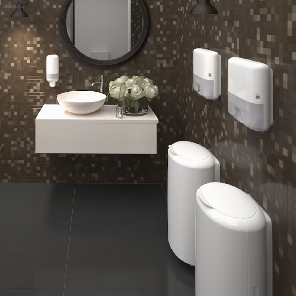 Un moderno bagno pubblico con una parete di piastrelle a mosaico marroni presenta uno specchio rotondo, un lavabo montato a parete con bacino bianco, dispenser di sapone, Tork Xpress® 552100 Mini Dispenser per Asciugamani Multifold Elevation H2 di TORK – Essity Professional Hygiene Germany GmbH e due cestini cilindrici bianchi. Un mazzo di fiori bianchi decora l'area del lavabo.