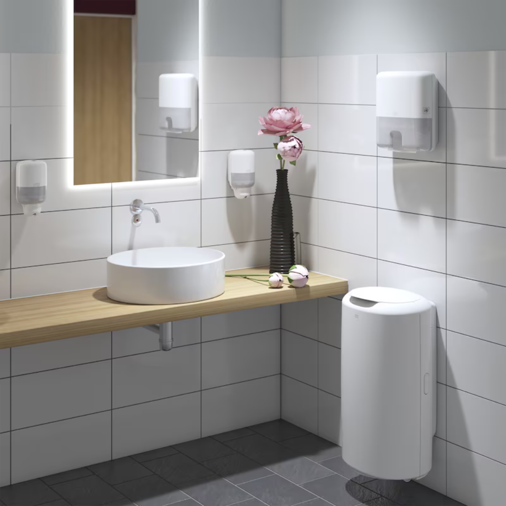 Un moderno bagno con un lavabo bianco rotondo su una mensola di legno, arricchito da uno specchio a parete con illuminazione. Il Tork Xpress® 552100 Mini Dispenser per Asciugamani Multifold Elevation H2 di TORK – Essity Professional Hygiene Germany GmbH dona eleganza alle pareti piastrellate bianche, insieme a un vaso nero pieno di fiori rosa. Sul pavimento piastrellato di grigio si trova un cestino bianco.