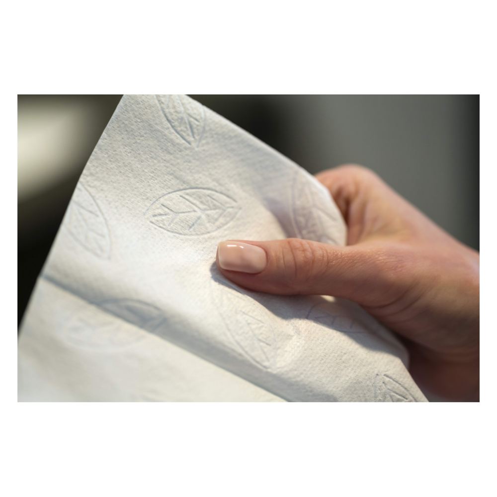 Eine Hand mit manikürten Nägeln hält ein Stück weißes Papiertuch. Das 2-lagige Papiertuch, das Tork 100278 extraweiche Zickzack-Handtücher Premium H3 2-lagig | Karton (15 Packungen) von TORK ähnelt, verfügt über ein geprägtes Design mit Blättern.