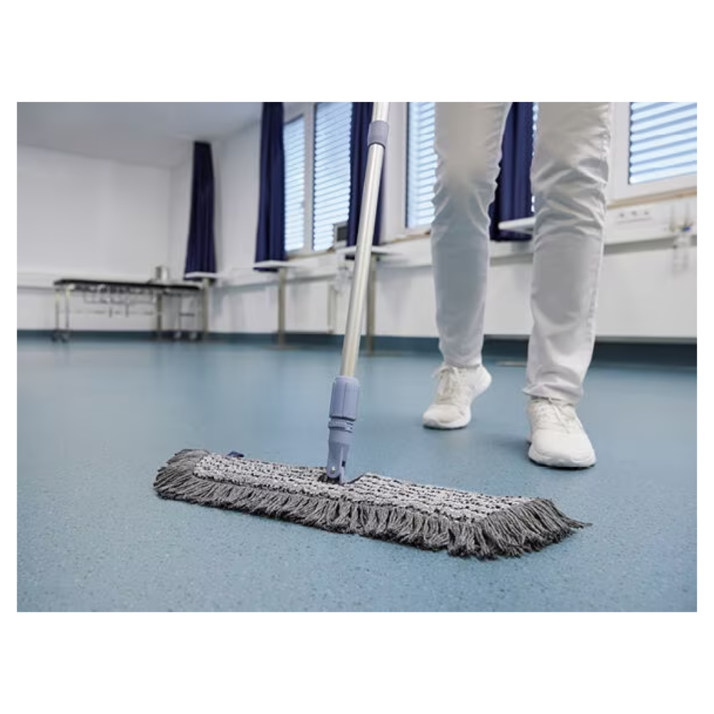 Una persona con scarpe e pantaloni bianchi pulisce un pavimento blu con un Vileda GmbH Professional Swep Duo r-MicroCombi Mop - 50 cm in una stanza con persiane chiuse e tende blu. Vicino ci sono un tavolo pieghevole e un radiatore sotto le finestre.