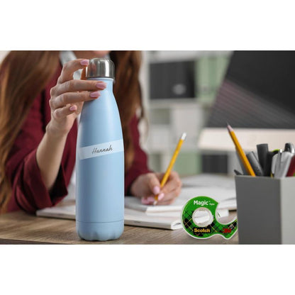 Una persona con una bottiglia d'acqua azzurra con la scritta "Hannah" è seduta a una scrivania con un quaderno aperto, matita e nastro adesivo invisibile Scotch® Magic™ della 3M Deutschland GmbH (2 rotoli, dispenser manuale), materiale da ufficio e scaffali sullo sfondo.