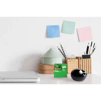 Una scrivania con un laptop chiuso, 3M Scotch™ Magic Tape, un dispenser da tavolo nero Scotch™ Karim Rashid di 3M Deutschland GmbH, bicchieri con penne e matite, scatole rotonde impilate e tre note adesive pastello su una parete bianca.