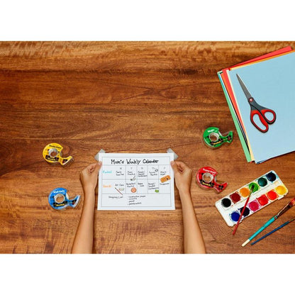 Una persona tiene un "Calendario Settimanale di Mamma" su un tavolo di legno con quattro dispenser di nastro adesivo, carta colorata, un set di acquerelli con pennello e una forbice di precisione rossa Scotch™ di 3M Deutschland GmbH.