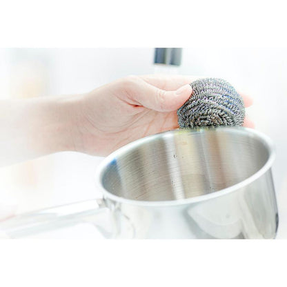 Una mano utilizza una spirale in acciaio inossidabile Scotch-Brite® di 3M Deutschland GmbH, 3 pezzi per confezione, per rimuovere lo sporco ostinato durante la pulizia di una pentola in acciaio inossidabile.