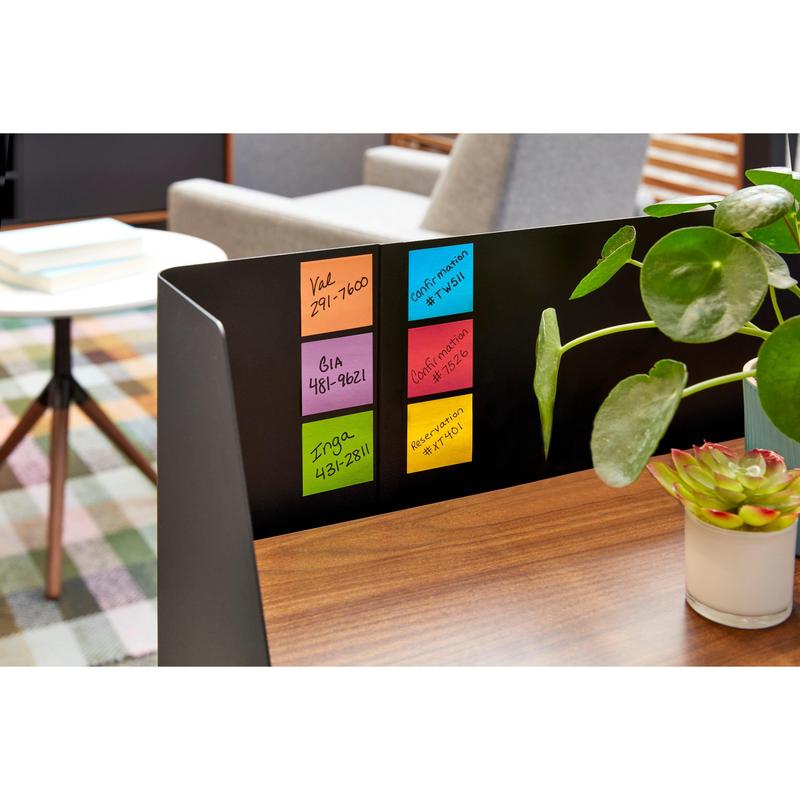Su una scrivania di legno ci sono cinque colorati Post-it® Super Sticky Z-Notes (76 x 76 mm, 90 fogli/blocco) della 3M Deutschland GmbH. Accanto a loro ci sono una pianta in vaso e una succulenta. Sullo sfondo: tavolo, sedie, tappeto a quadri.