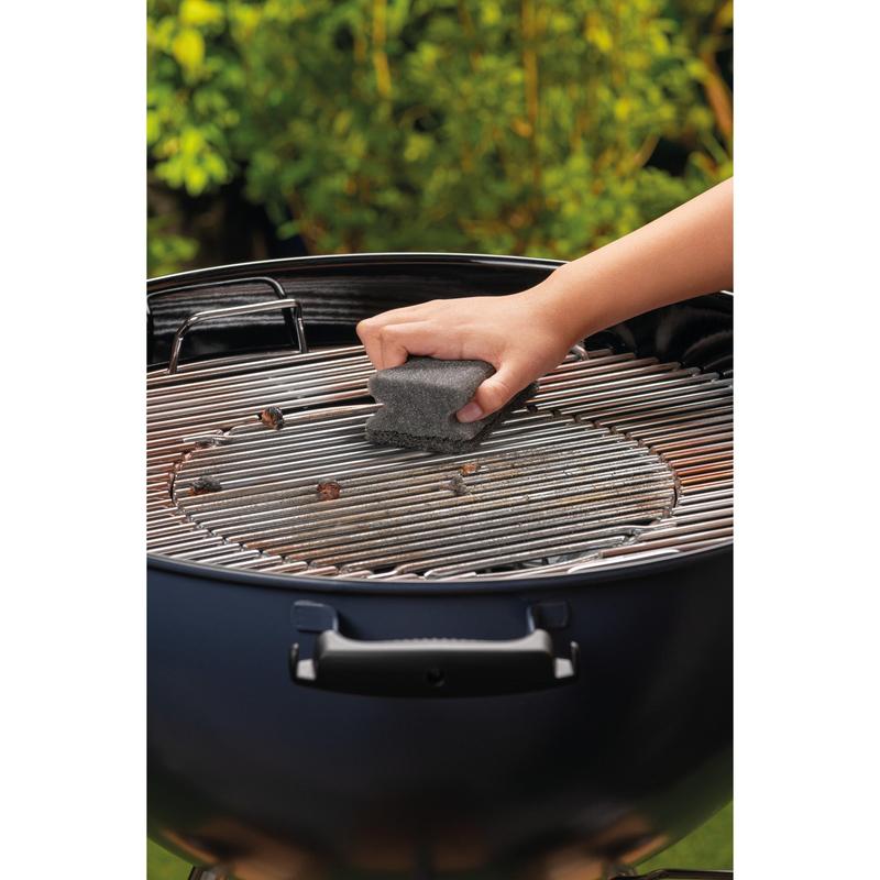 Una mano pulisce la griglia metallica di un barbecue a carbone rotondo con una spugna Scotch-Brite® Grill (1 pezzo per confezione) della 3M Deutschland GmbH. Fogliame verde è sfocato sullo sfondo.