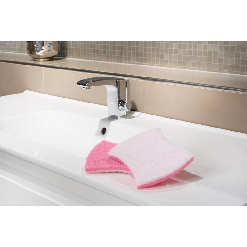 Un lavabo bianco con un rubinetto cromato mostra la spugna per la pulizia del bagno Scotch-Brite® Bath XXL di 3M Deutschland GmbH, rosa da un lato e bianca dall'altro, ideale per rimuovere i depositi di calcare. Sullo sfondo appaiono piastrelle beige e marrone chiaro.