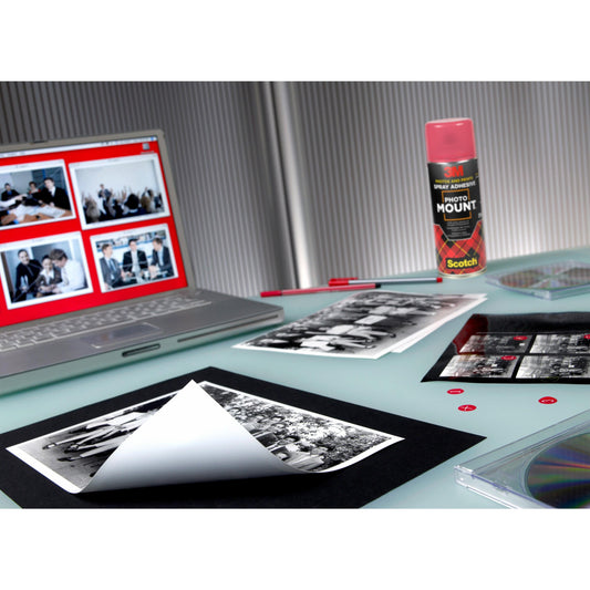 Auf einem Schreibtisch stehen Schwarz-Weiß-Fotos, ein Laptop mit Foto-Miniaturansichten, ein roter Stift, CDs, kleine rote Aufkleber und eine Dose 3M™ Scotch® PhotoMount™ Klebespray (400 ml) der 3M Deutschland GmbH auf einer hellen Fläche.